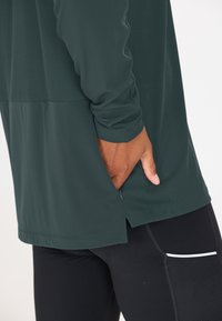 Dunkelgrünes langärmliges Sportoberteil mit strukturierten Einsätzen, seitlichem Schlitzsaum und kontrastierendem glattem unteren Bereich, kombiniert mit schwarzen eng anliegenden Leggings.