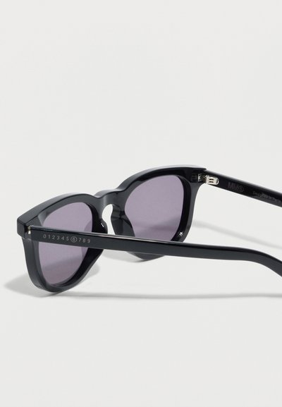 Μαύρα γυαλιά ηλίου από οξικούς με φακούς στρογγυλής μορφής με μοβ απόχρωση και λεπτούς βραχίονες, τοποθετημένα σε λευκή επιφάνεια.