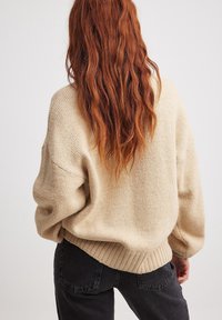 Pull en tricot beige oversize avec des poignets et un ourlet côtelés. Texture douce avec une coupe décontractée, associé à un jean sombre. Les cheveux tombent dans le dos.