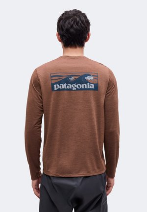 Mand iført brun langærmet skjorte med blå Patagonia-logo med havbølger, set bagfra mod en ensfarvet baggrund.
