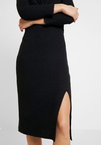 Svart stickad klänning med ribbat mönster, långa ärmar och en sidoklyvning, med en figurnära silhuett som framhäver kroppens form.