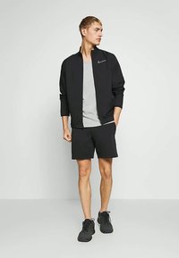 Jauns vīrietis stāv, tērpies melnā Nike jakā, pelēkā T-kreklā, melnās šortos un melnās sporta apavās, pret vienkāršu gaišu fona.