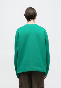 Πράσινο φούτερ από απαλό ύφασμα, με άνετη εφαρμογή, στρογγυλή λαιμόκοψη και κατεβασμένους ραφές ώμων. Φορεμένο πάνω από σκούρο παντελόνι cargo.