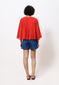 Rotes, leichtes Cape-Top mit geraffter Textur an den Schultern, kombiniert mit blauen Denim-Shorts. Modell trägt rote Sandalen, von hinten betrachtet.