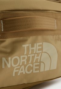 Μπεζ τσάντα The North Face με ορατό υφαντό λουρί και μεγάλο λευκό λογότυπο της μάρκας τυπωμένο στην επιφάνεια του υφάσματος.