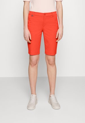 Person iført lyse orange knælange shorts, lyserød top og hvide sneakers, der står mod en ensfarvet lys baggrund.