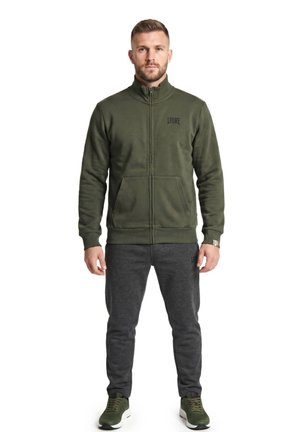Un homme porte une veste zippée vert olive, un pantalon gris foncé et des baskets vertes sur fond blanc.
