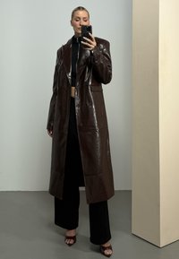 Γυναίκα που βγάζει selfie μπροστά στον καθρέφτη φοράει μακρύ σκούρο καφέ δερμάτινο παλτό πάνω από μαύρο παντελόνι, μαύρο κοντό τοπ και ανοιχτά πέδιλα σε εσωτερικό χώρο.
