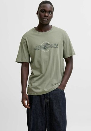 Ung mand med en neutral udtryk iført en grøn "Jack & Jones" t-shirt og mørkeblå jeans, med den ene hånd i lommen.