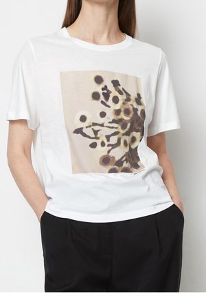 Frau trägt weißes T-Shirt mit kurzen Ärmeln und abstraktem braun-beigeem Kreisdesign, kombiniert mit schwarzer Hose, Hände in den Taschen.