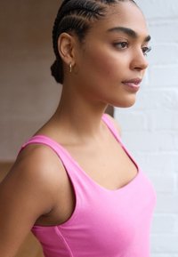 Top rosa attillato con scollatura rotonda e spalline larghe, realizzato in tessuto morbido. La modella ha capelli intrecciati e piccoli orecchini a cerchio.