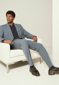 Helles graues Anzug mit einer taillierten Blazerjacke und geradem Hosenbein, getragen über einem schwarzen V-Ausschnitt-Shirt. Schwarze Slipper und marineblaue Socken runden den Look ab.