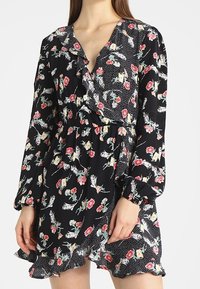 Černé květinové šaty s dlouhými rukávy, výstřihem do V, volány a puntíkovaným vzorem na spodním lemu. Obsahují barevný květinový potisk na tmavém pozadí.