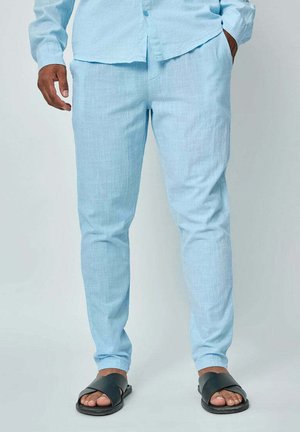 Homme portant un pantalon en lin bleu clair et une chemise assortie avec des sandales noires, debout, une main dans la poche, devant un fond uni.