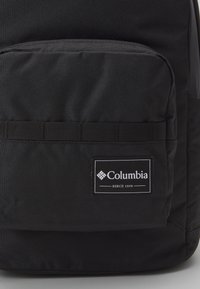 Czarny plecak z trwałego materiału, z przednią kieszenią, poziomym detalem z taśmy i białą łatką z logo z napisem "Columbia".