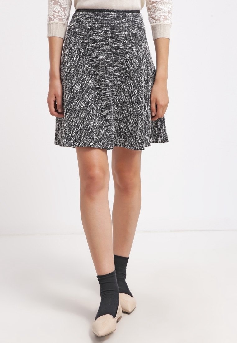 Femme portant une jupe trapèze texturée gris et blanc, des chaussettes noires, des ballerines beiges et un haut crème à manches en dentelle, debout sur un fond blanc.