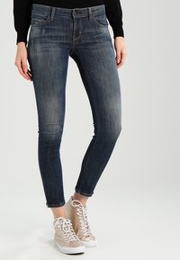 Slim fit tmavě modré džíny s vybledlou texturou, které mají dvě přední kapsy a zipový uzávěr, doplněné béžovými vysokými teniskami.