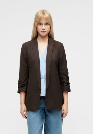 Jeune femme aux cheveux blonds raides portant un blazer noir par-dessus un haut bleu clair et un jean bleu, debout devant un fond clair uni.