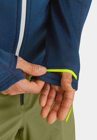 Navy athletic jacket with a white zipper and neon yellow accent, featuring thumb holes and a close-fitting design on a green background.