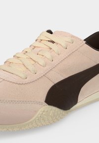 Gros plan d'une basket rose clair avec des lacets crème et une bande latérale marron foncé sur une semelle beige texturée.