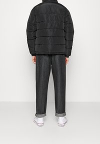 Persona che indossa un giubbotto puffo nero, pantaloni grigi a righe con risvolti e sneakers bianche Converse All Star, in piedi contro uno sfondo bianco.