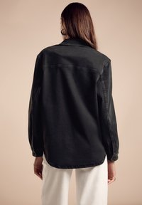 Svart denimskjorta med långa ärmar, knappmanschetter, spetsig krage och rak skärning, bärs över vita byxor och visar en jämn textur.