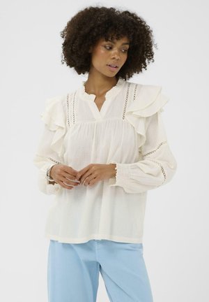 Blusa color crema con mangas con volantes, tela texturizada y pequeños detalles de encaje, combinada con pantalones azul claro. Puños largos y elásticos.