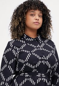 Abito nero con motivo a zigzag bianco, caratterizzato da un colletto, chiusura frontale con bottoni e vita con cintura. Tessuto dalla texture liscia.