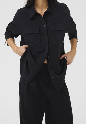 Personne portant une chemise noire ample boutonnée avec des poches avant et un pantalon large assorti, les mains posées sur les hanches.