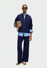 Navy zip-up jacket over a light blue shirt, paired with navy wide-leg pants and beige shoes. Accessories include a leopard print handbag.