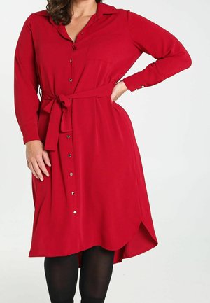 Femme portant une robe rouge à manches longues avec boutons et une ceinture à la taille, debout avec une main sur la hanche et un legging noir en dessous.