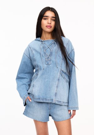 Denim pulover s kapuco, z vezanjem spredaj, dvema sprednjima žepoma, v svetlo modri barvi. Ujemajoče kratke hlače iz denima z raztrgano obrobo.