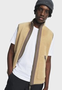 Beige Fleece-Weste mit hohem Kragen, Reißverschluss und braunen Akzenten. Verfügt über Seitentaschen und eine texturierte Außenseite. Getragen über einem weißen T-Shirt.