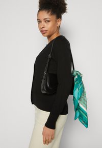 Zwarte gebreide cardigan met een getailleerde pasvorm, gecombineerd met een kleine zwarte handtas en een turkooise zijden sjaal met geometrische patronen.