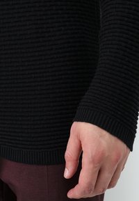 Pull noir en maille avec un motif côtelé texturé, doté de manches longues et d'un poignet ajusté, porté sur un pantalon bordeaux.