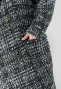 Manteau en mélange de laine dans un motif pied-de-poule gris et noir avec de grands boutons noirs et une poche latérale, arborant une étiquette de marque sur la manche.