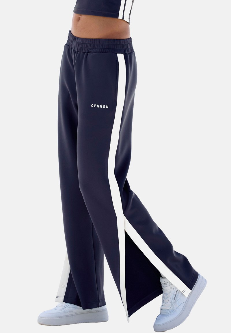 Pantalones atléticos en color navy con franjas laterales blancas, fabricados en una tela suave, que presentan un corte relajado y aberturas en el dobladillo para mayor estilo. Logo en el muslo.