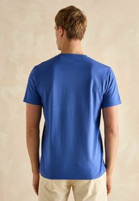 Niebieska bawełniana koszulka z krótkim rękawem i okrągłym dekoltem. Gładka faktura i luźny krój, pokazana z tyłu na neutralnym tle.