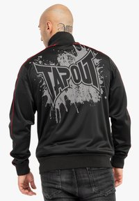 Chaqueta negra con acentos rojos, que presenta un gran gráfico gris "TAPOUT" en la parte posterior. Textura suave y corte atlético con cuello con cremallera.