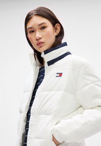 Balta pūkų striukė su aukštu apykakle, turinti tamsiai mėlynų akcentų ir logotipu kairėje krūtinės pusėje. Lygi tekstūra ir siūti raštai.