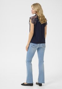 Marinblå topp med spetsdetaljer på axlarna, i kombination med ljusblå utsvängda jeans och svarta platta skor, sedd bakifrån.