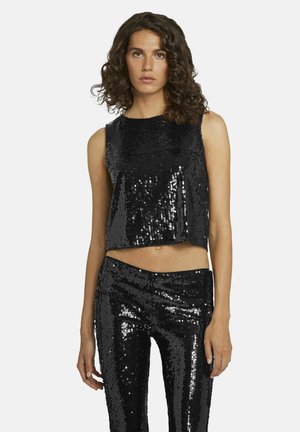 Vrouw met krullend haar die een zwart mouwloos topje met pailletten en bijpassende hooggetailleerde paillettenbroek draagt, staand tegen een witte achtergrond.