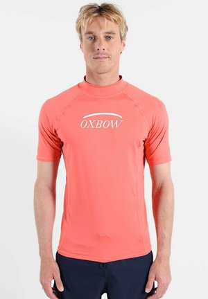 Homme blond portant un rash guard corail à manches courtes avec le logo "OXBOW", debout sur un fond blanc uni.