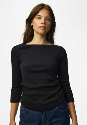Jeune femme aux cheveux bruns portant un haut noir à manches longues avec un col bateau et un jean bleu sur un fond blanc uni.