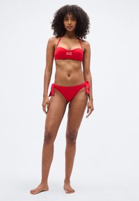 Σετ κόκκινου μπικίνι με τοπ bandeau που διαθέτει λεπτές τιράντες και λογότυπο. Το κάτω μέρος έχει χαμηλή μέση και δένει στα πλαϊνά. Ομαλή υφή.
