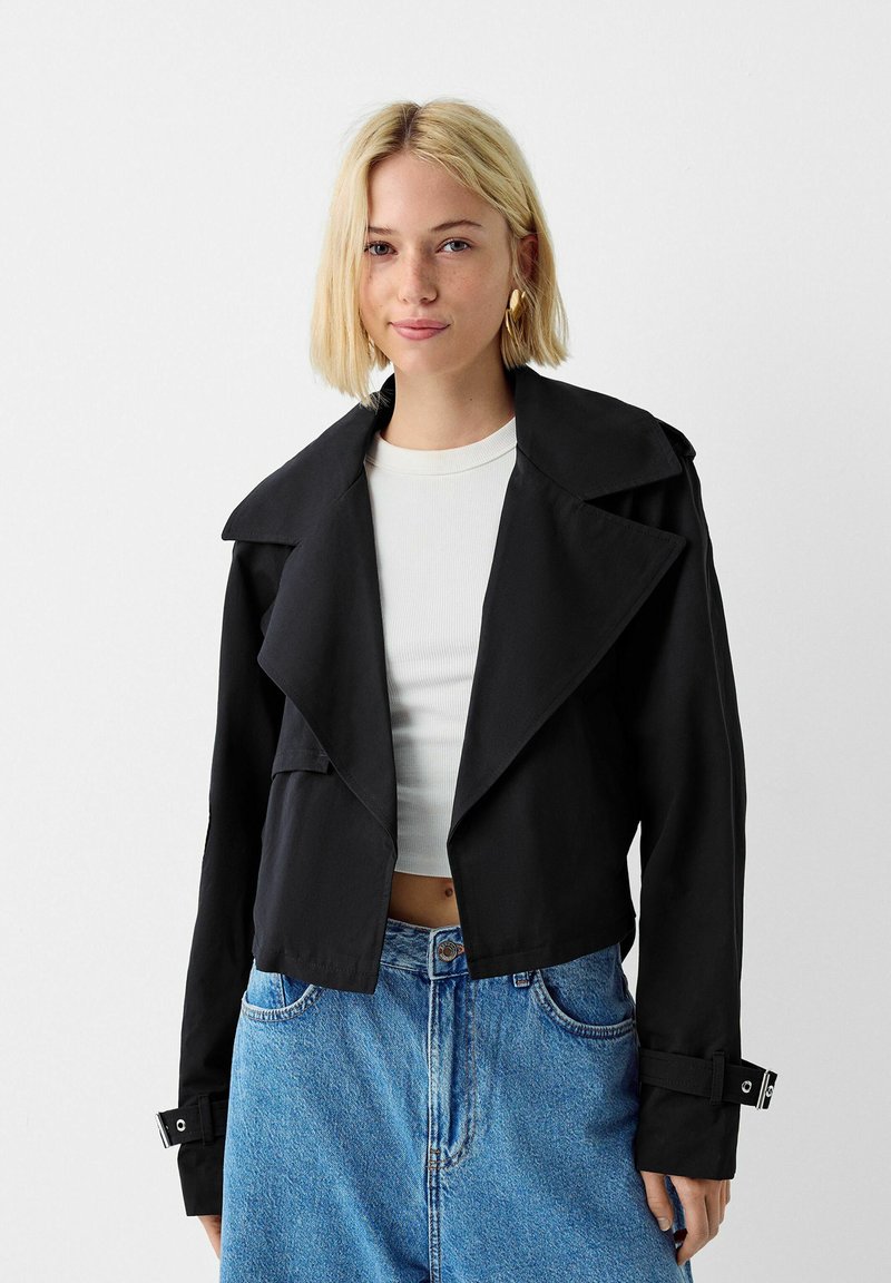 Giacca nera corta con colletto ampio e tasche frontali, realizzata in un tessuto liscio. Abbinata a una maglietta bianca e jeans in denim blu a vita alta.