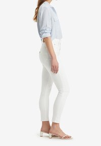Jeans ajustados blancos combinados con una camisa de rayas azul claro. El conjunto presenta mangas enrolladas y sandalias beiges de tacón cuadrado.