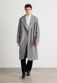 Grå uld frakke med dobbeltradet design, brede revers og forlommer, båret over en hvid skjorte og sorte bukser.