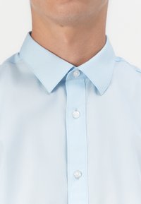 Gros plan d'une personne portant une chemise bleu clair à boutons avec un col pointu, montrant trois boutons blancs attachés sur le devant.
