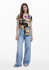 Bunt bedrucktes T-Shirt mit abstrakten Mustern, kombiniert mit hellblauen weit geschnittenen Jeans. Das Model trägt eine kleine geflochtene Tasche mit einem Trageriemen.